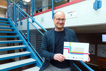 Maarten Willenbrink, Leiter der Maximilian-Kolbe-Gesamtschule, hält das Schild mit der Aufschrift Euregioprofilschule hoch, das es im Zuge der Zertifizierung gab und das demnächst an einer Wand im Schulgebäude hängen wird. Foto: Katja Niemeyer